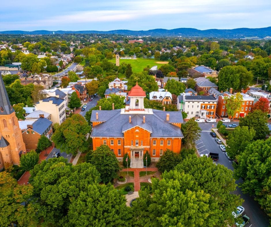 Aerial view of a historic red brick government building with a white bell tower surrounded by lush green trees and a small town cityscape.